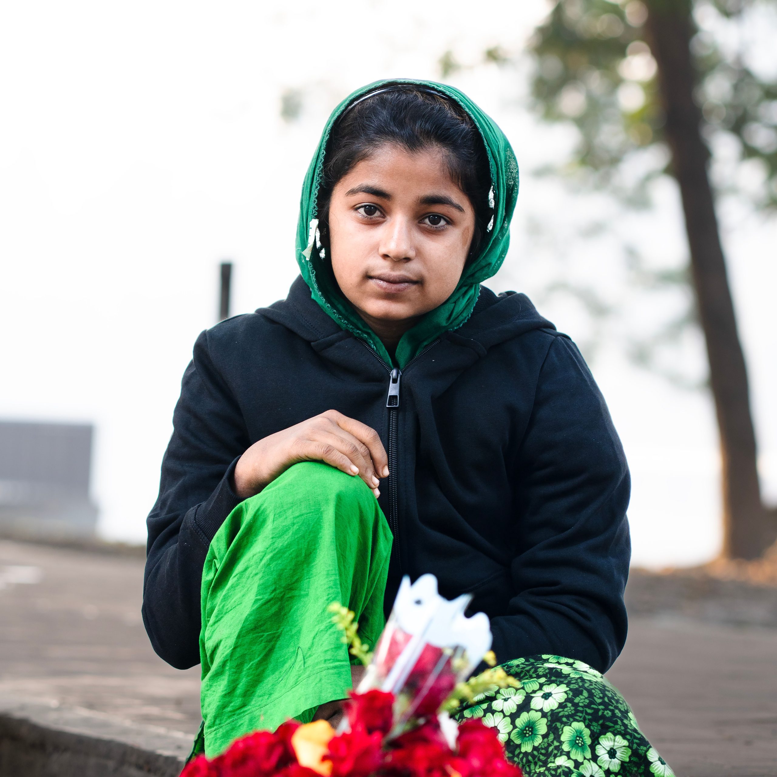 A young girl in a green headscarf and black jacket sits outdoors, holding a bouquet of red flowers.