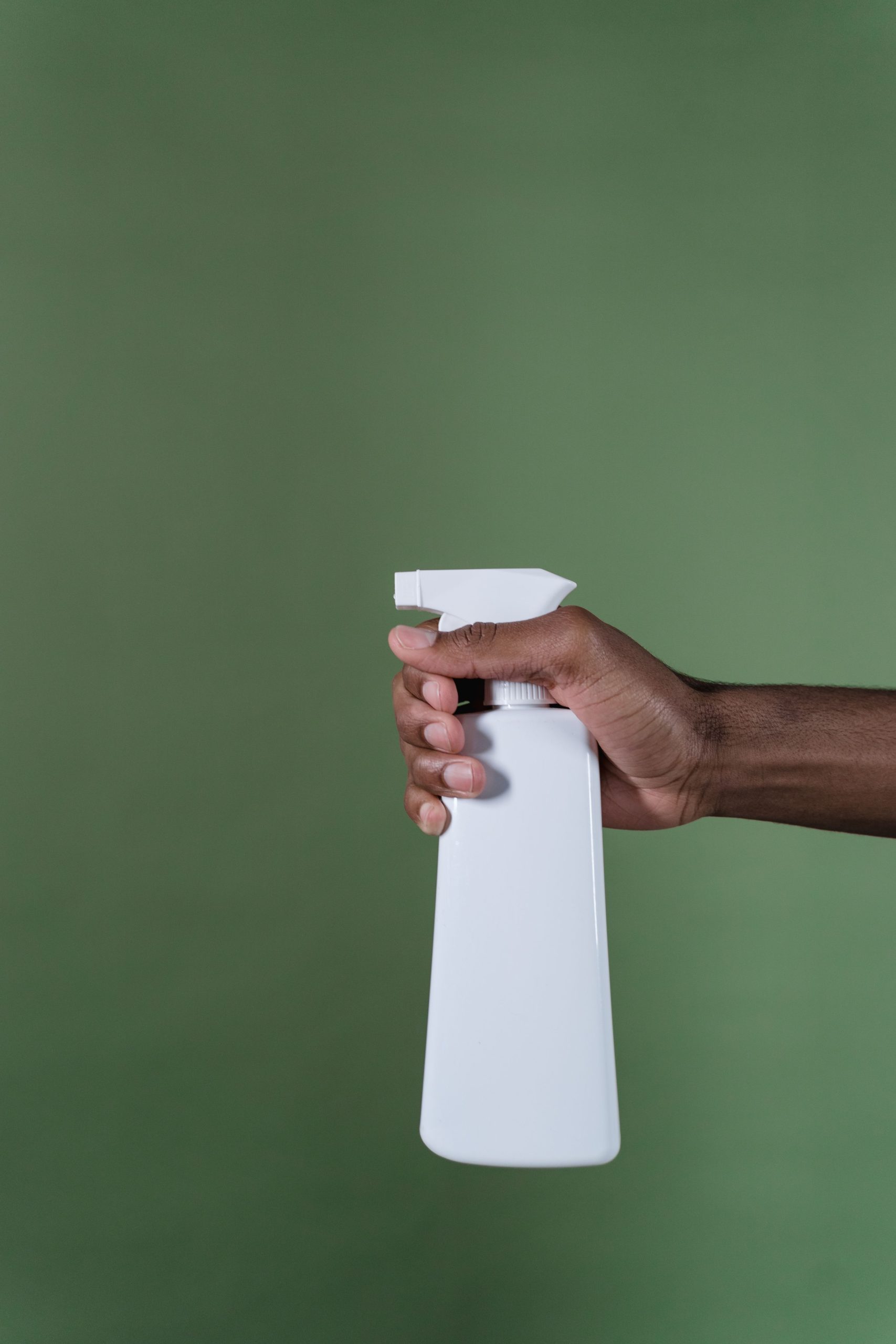 A close-up image of a hand holding a white spray bottle against a plain green background.